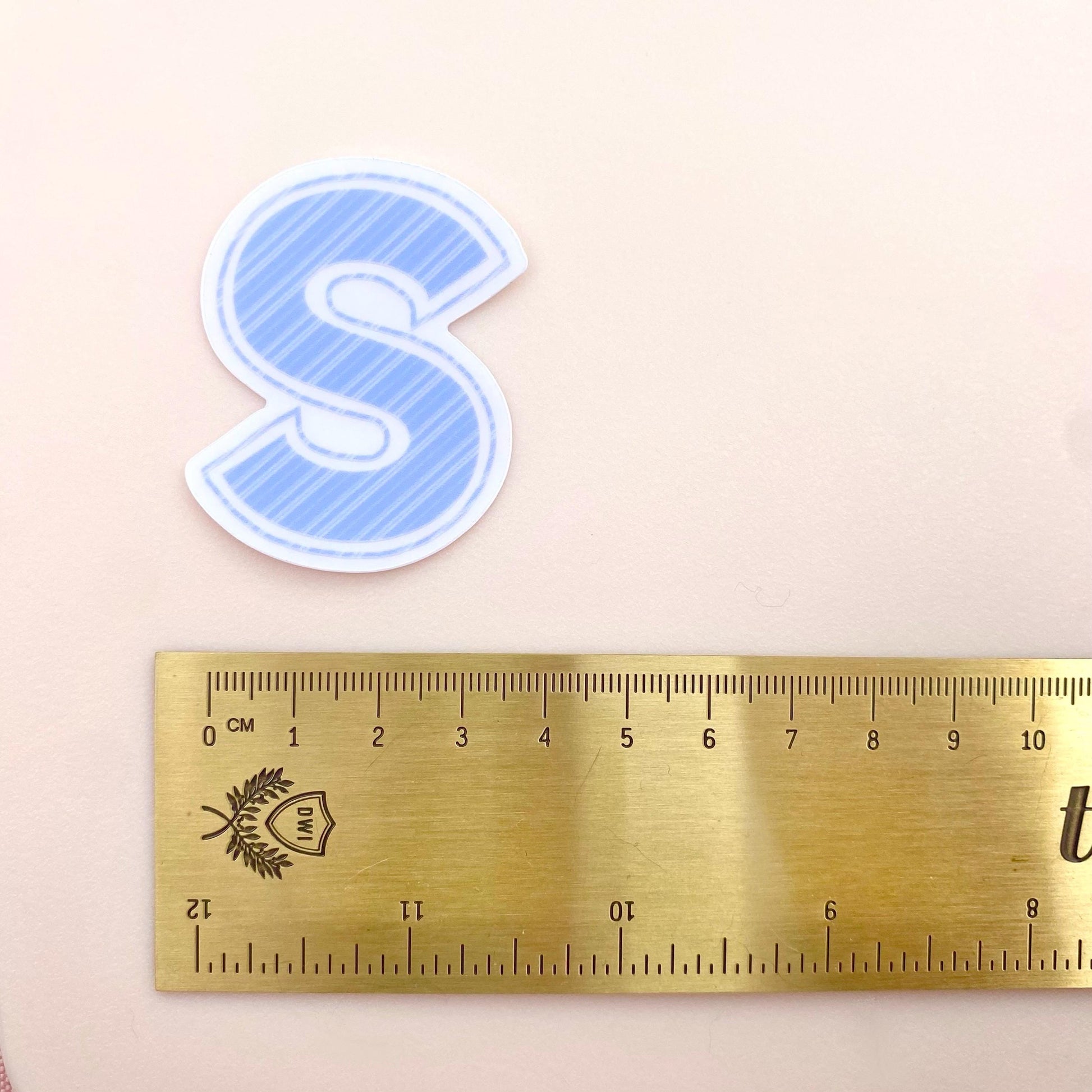 Blue striped letter 'S' sticker next to a gold ruler on a beige background