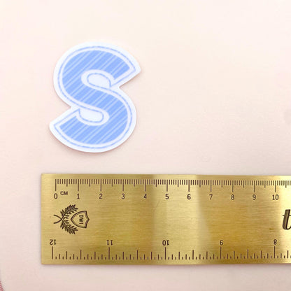 Blue striped letter 'S' sticker next to a gold ruler on a beige background