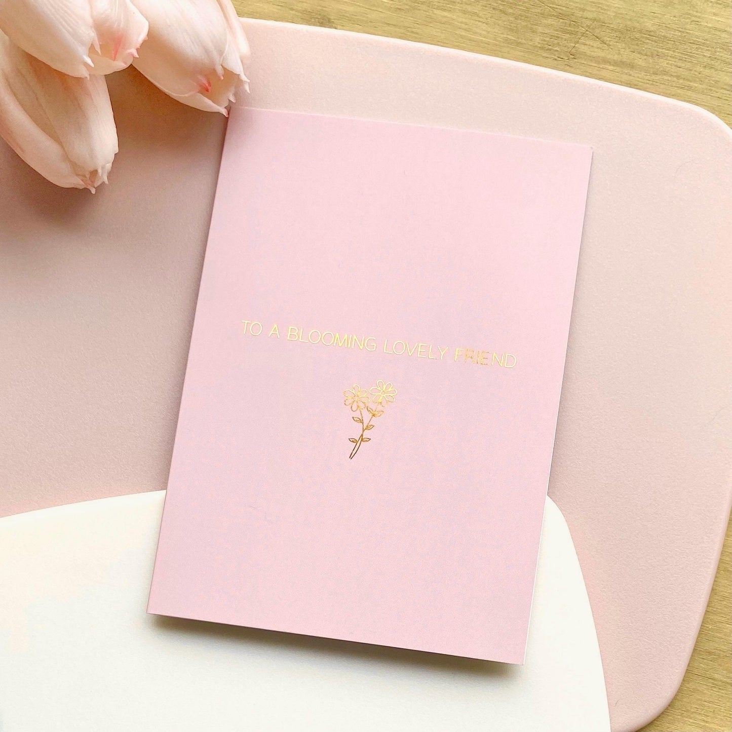 Pink greeting card with gold text and floral design on a light pink surface with tulips.