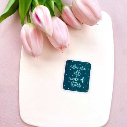 A blue sticker with white glowing text 'We are all made of stars' on a cream background with pink tulips.