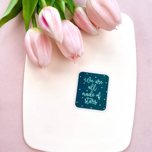 A blue sticker with white glowing text 'We are all made of stars' on a cream background with pink tulips.