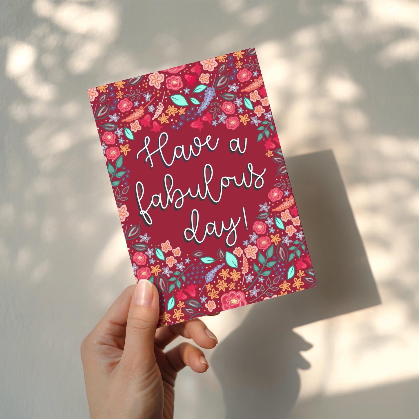 Hand holding a dark pink card with floral design and 'Have a fabulous day!' text.