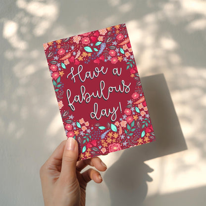 Hand holding a dark pink card with floral design and 'Have a fabulous day!' text.