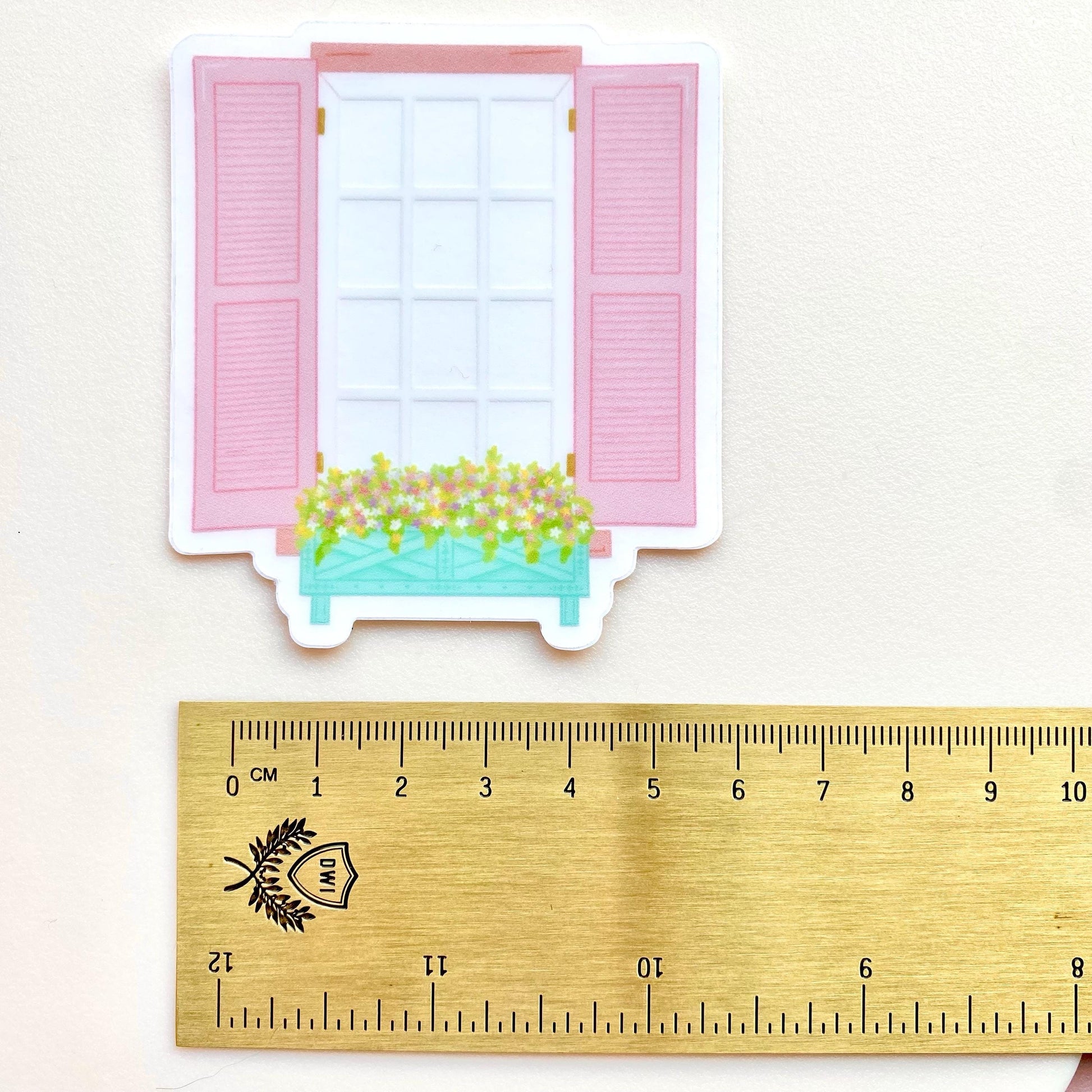 Transparent sticker of a window with pink shutters and a flower box on a white background with a gold ruler for scale.