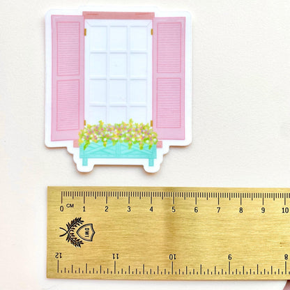 Transparent sticker of a window with pink shutters and a flower box on a white background with a gold ruler for scale.