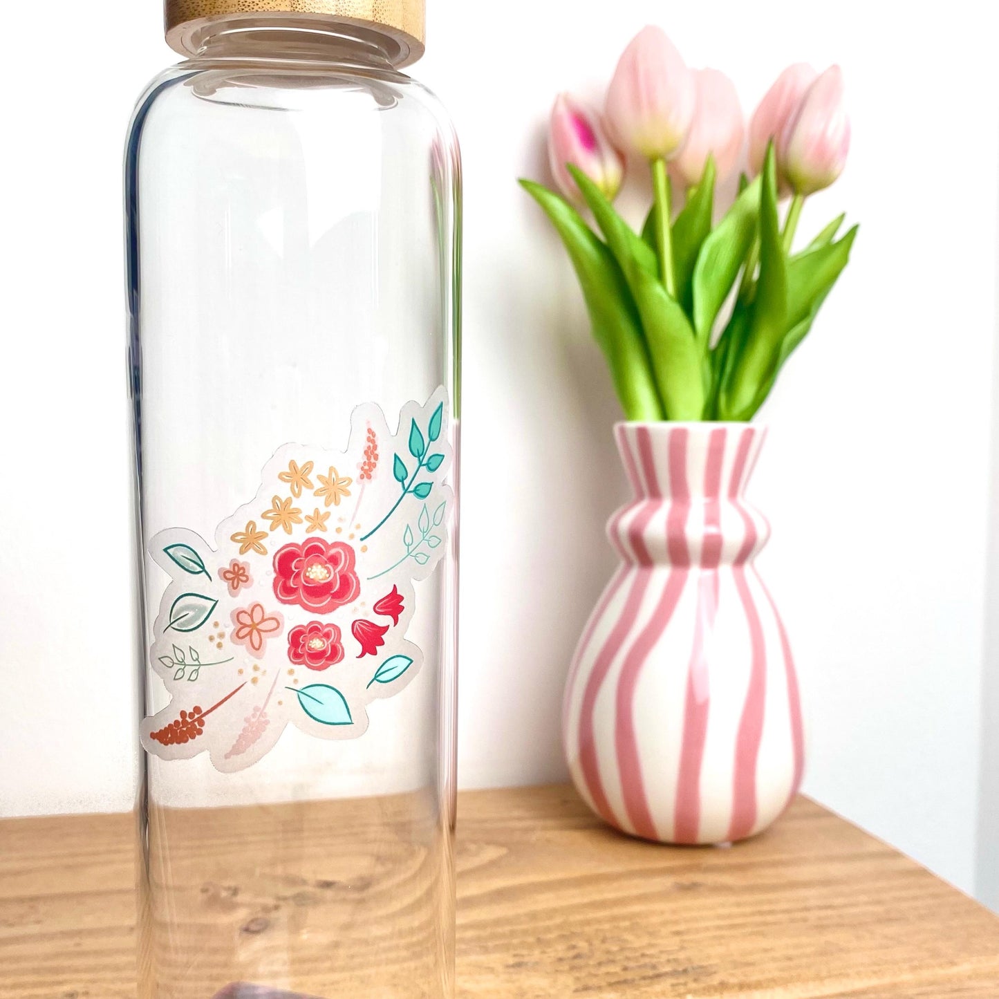 Clear glass bottle with floral sticker next to a pink and white striped vase with tulips on a wooden surface.