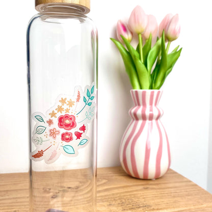 Clear glass bottle with floral sticker next to a pink and white striped vase with tulips on a wooden surface.