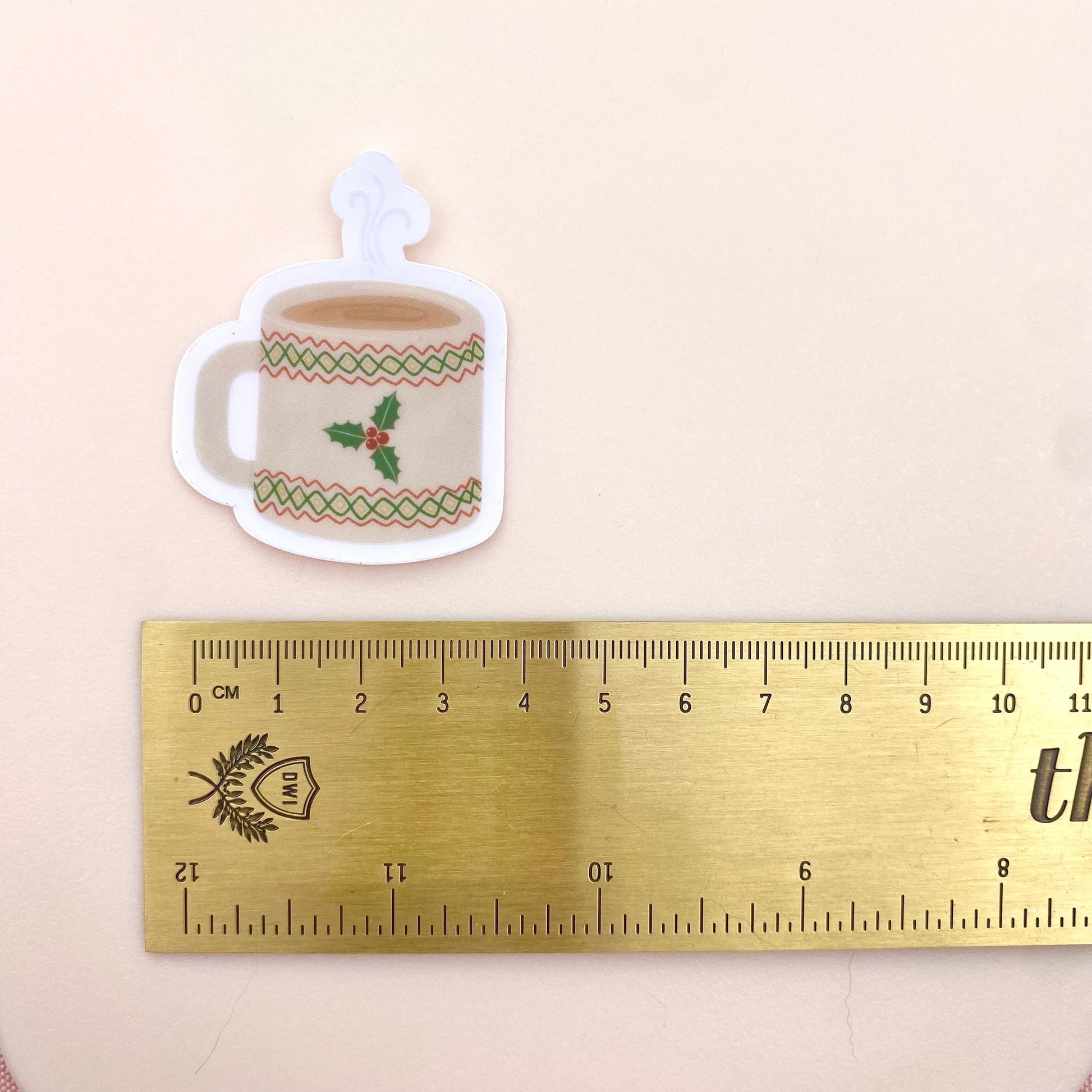 Sticker of a Christmas-themed mug with holly design next to a ruler for scale on a beige background