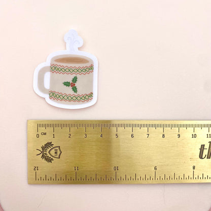 Sticker of a Christmas-themed mug with holly design next to a ruler for scale on a beige background
