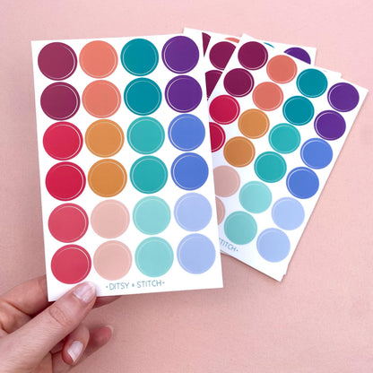 A sheet of colourful dot stickers held by a hand on a pink background