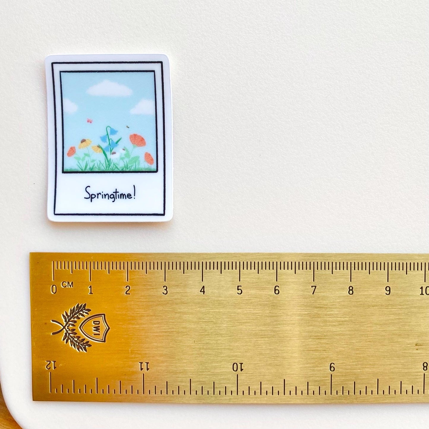 Clear sticker with a polaroid design showing a spring scene with the text 'Springtime!' shown with a gold ruler for scale