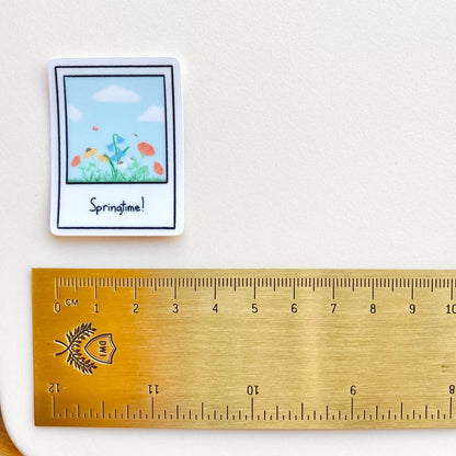 Clear sticker with a polaroid design showing a spring scene with the text 'Springtime!' shown with a gold ruler for scale