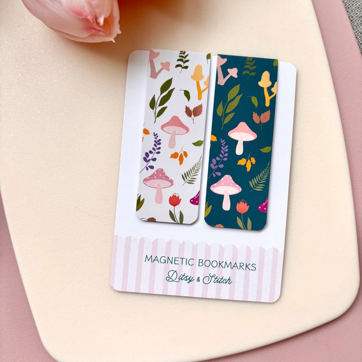 Two decorative magnetic bookmarks with autumn mushroom and botanical illustrations are displayed on a branded card that reads 'MAGNETIC BOOKMARKS Ditty & Stitch.' The bookmarks are photographed on a soft cream surface with a pale pink flower prop in the corner.
