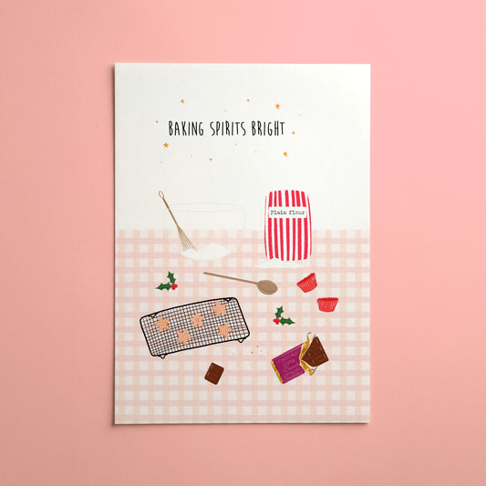 A rectangular baking-themed Christmas art print with the phrase 'BAKING SPIRITS BRIGHT' at the top, shown against a solid pink background. The print features a white upper section and a pale pink gingham lower section with illustrated baking items including a jar labeled 'Plain Flour', a spoon, a cooling rack with cupcakes, and small red cupcakes cases and holly.