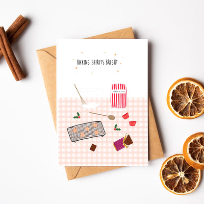 Greeting card with Christmas baking illustrations in pink gingham and 'Baking Spirits Bright' text, surrounded by cinnamon sticks and dried oranges on a white background, with a brown kraft card.