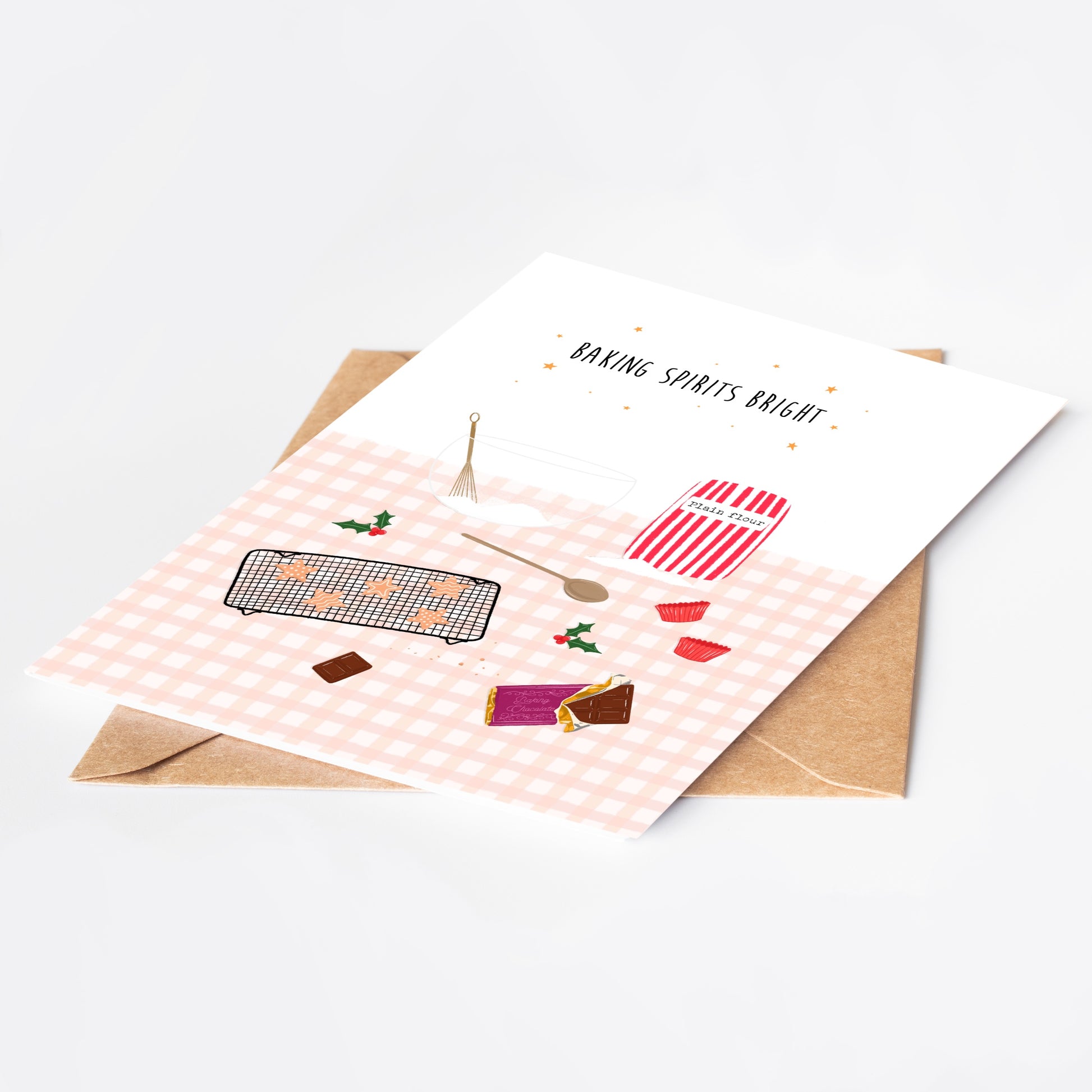 Greeting card with Christmas baking-themed illustrations on a pink gingham background, placed on a white surface with a brown kraft envelope.