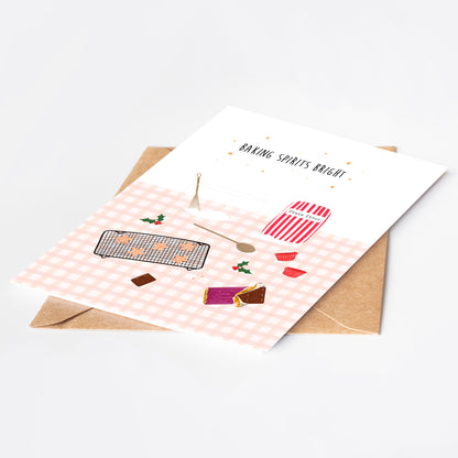 Greeting card with Christmas baking-themed illustrations on a pink gingham background, placed on a white surface with a brown kraft envelope.