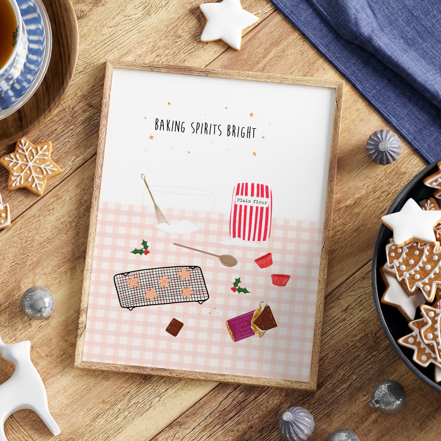 Framed print with Christmas baking-themed design on a wooden table with star cookies, a bowl, whisk, wooden spoon, bag of flour and chocolate.