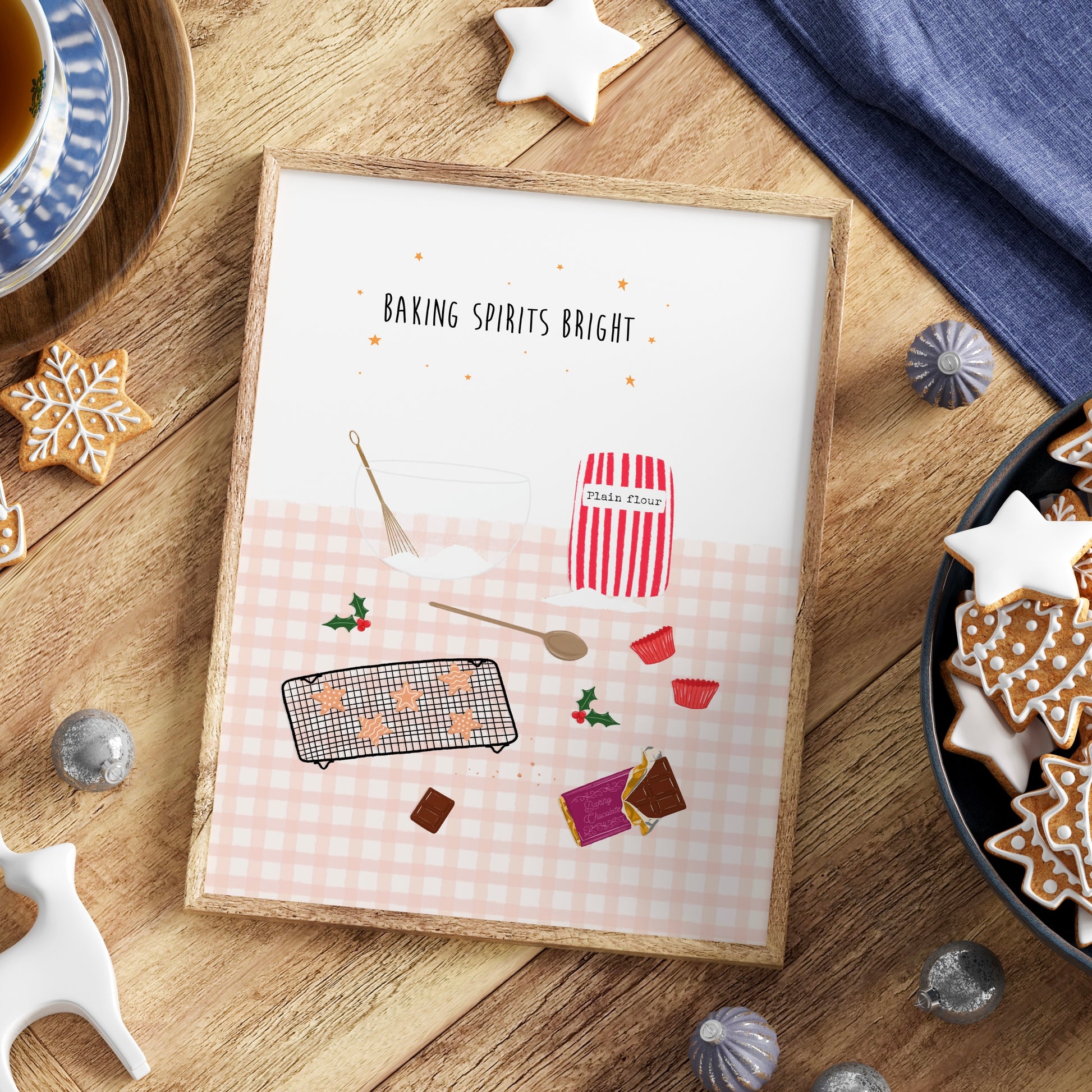 Framed print with Christmas baking-themed design on a wooden table with star cookies, a bowl, whisk, wooden spoon, bag of flour and chocolate.
