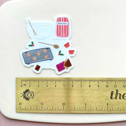 Sticker with festive baking tools and star shaped cookies and chocolate on a white surface with a ruler for scale.