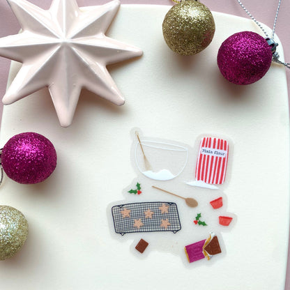 A sticker with an illustration of christmas baking set with a red and white striped flour bag, wooden spoon, mixing bowl and whisk, and star shaped cookies, with holly on a white background with pink and gold glittery baubles.