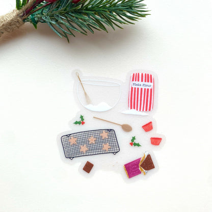 A sticker with an illustration of christmas baking set with a red and white striped flour bag, wooden spoon, mixing bowl and whisk, chocolate and star shaped cookies, with holly on a white background.