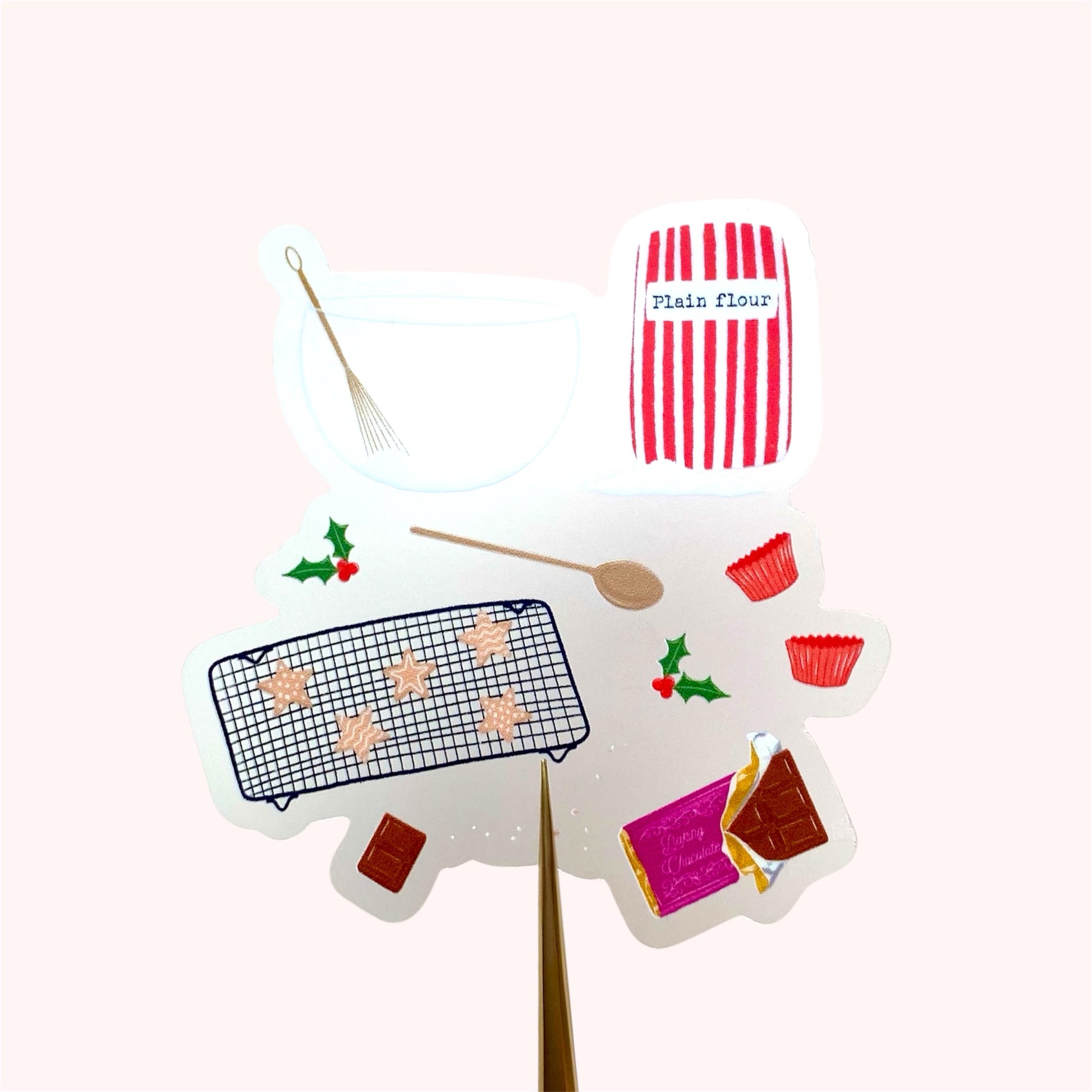 A sticker with an illustration of christmas baking set with a red and white striped flour bag, wooden spoon, mixing bowl and whisk, chocolate and star shaped cookies, held up against a pink background.