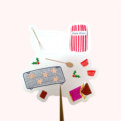 A sticker with an illustration of christmas baking set with a red and white striped flour bag, wooden spoon, mixing bowl and whisk, chocolate and star shaped cookies, held up against a pink background.