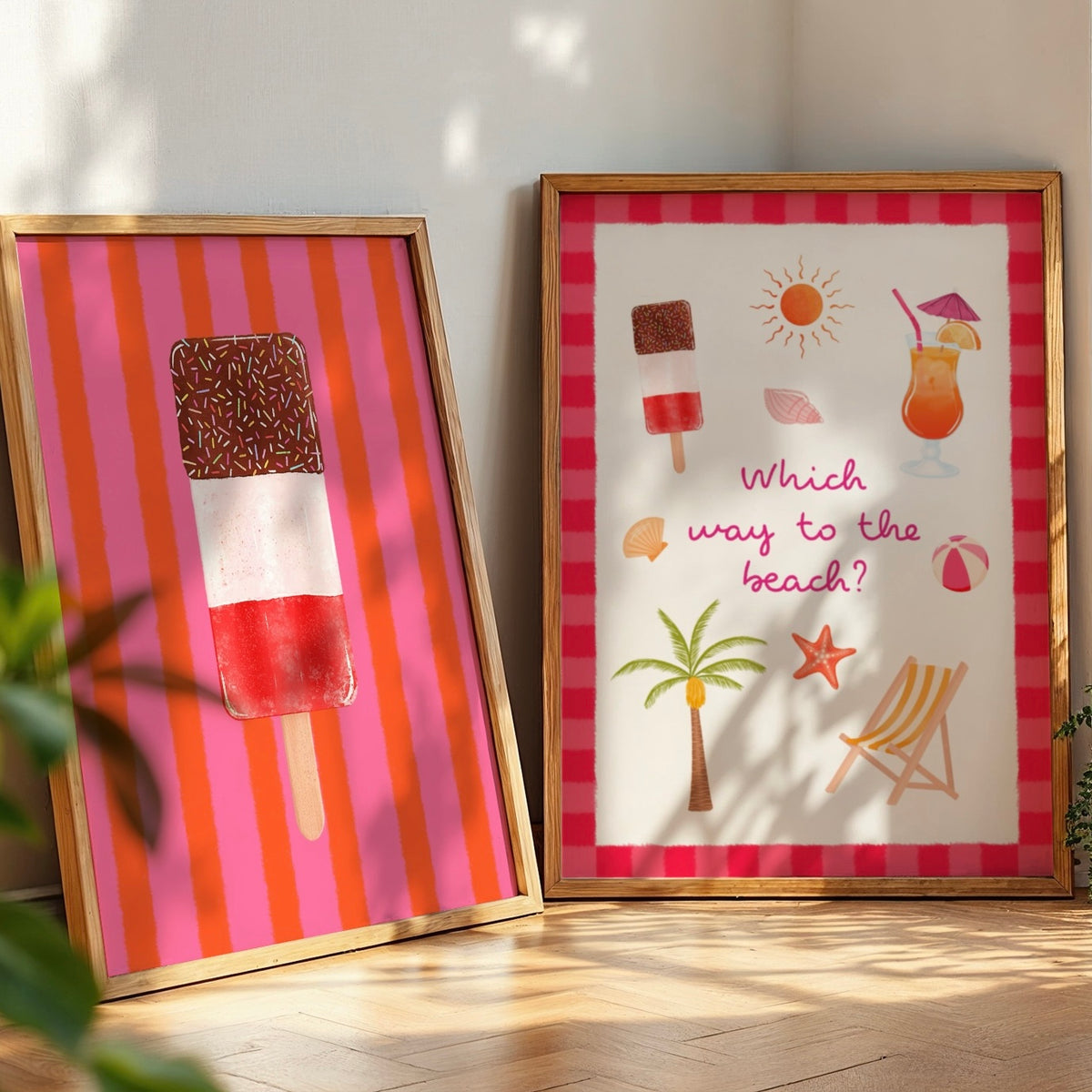 Two framed summer-themed art prints leaning against a wall on a wooden floor. The left print has pink and orange vertical stripes with an ice lolly illustration and the right has beach themed illustrations.