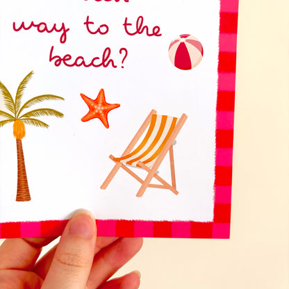 A close-up of a hand holding a white beach-themed art print with red text reading 'way to the beach?', decorated with a palm tree, starfish, beach chair, and beach ball, plus a red and pink gingham border.