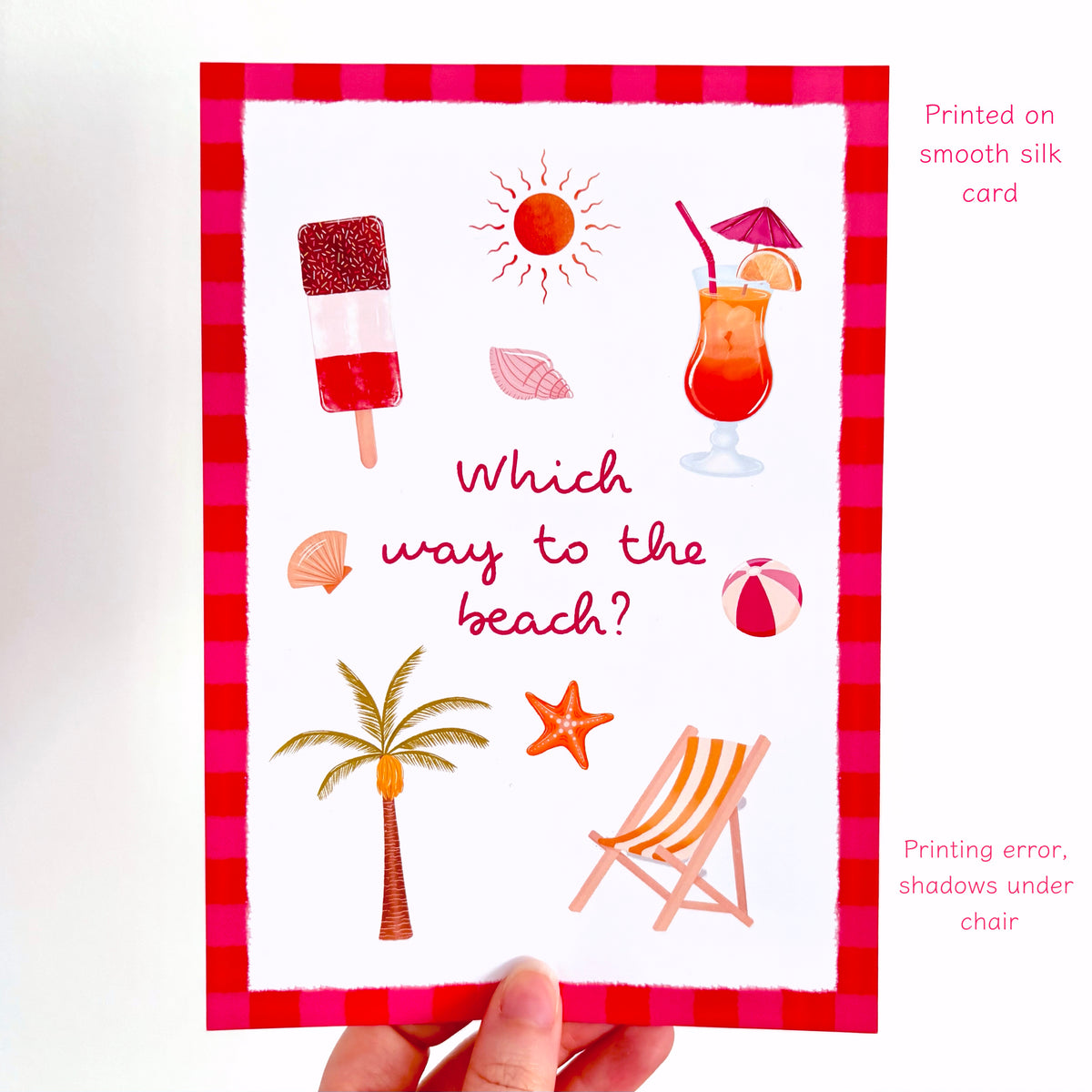 A hand holds a white silk greeting card featuring a red and pink gingham border and summer-themed illustrations including an ice lolly, sun, cocktail, beach umbrellas, palm tree, starfish, and deck chair. Text on the right notes it is printed on a smooth silk card and mentions a printing error and shadow under the chair.