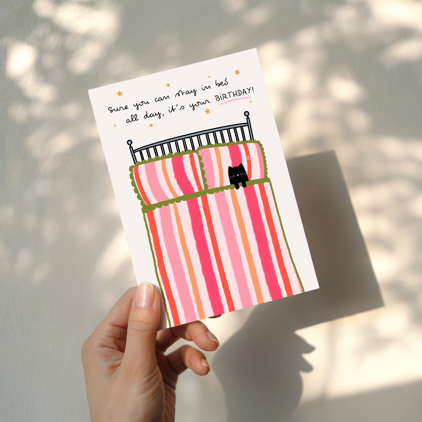 Hand holding a birthday card with a cat in bed illustration and humorous text.