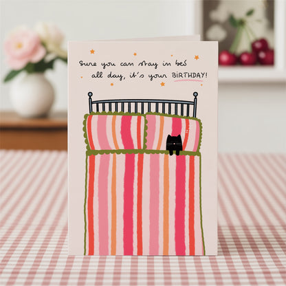 Birthday card with a cat in bed illustration with a pink, green and orange striped duvet, on a pink gingham tablecloth.