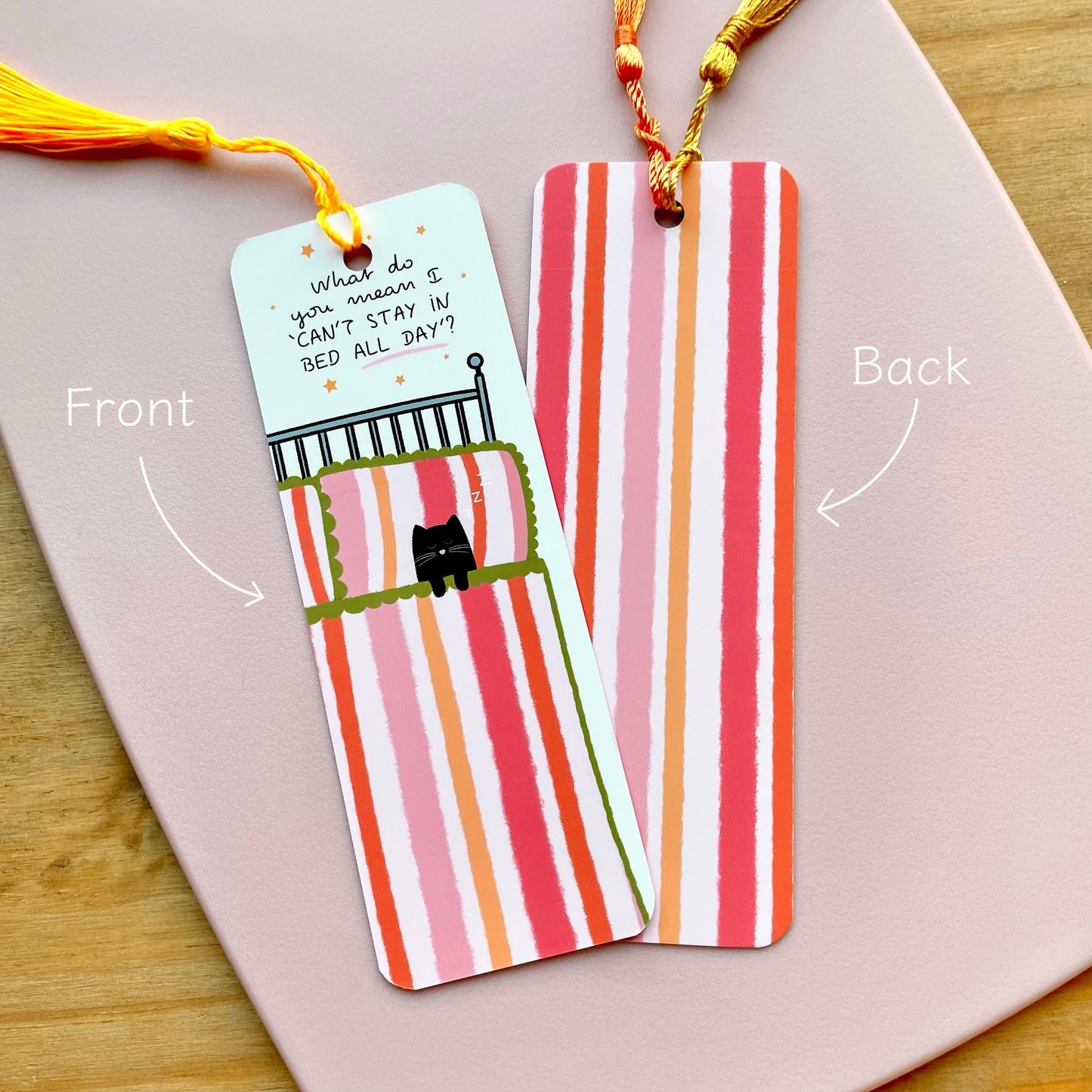 Two identical pink striped bookmarks, one with a cat illustration and text on the front, the other showing the back with vertical stripes in pink and orange, on a pink surface.