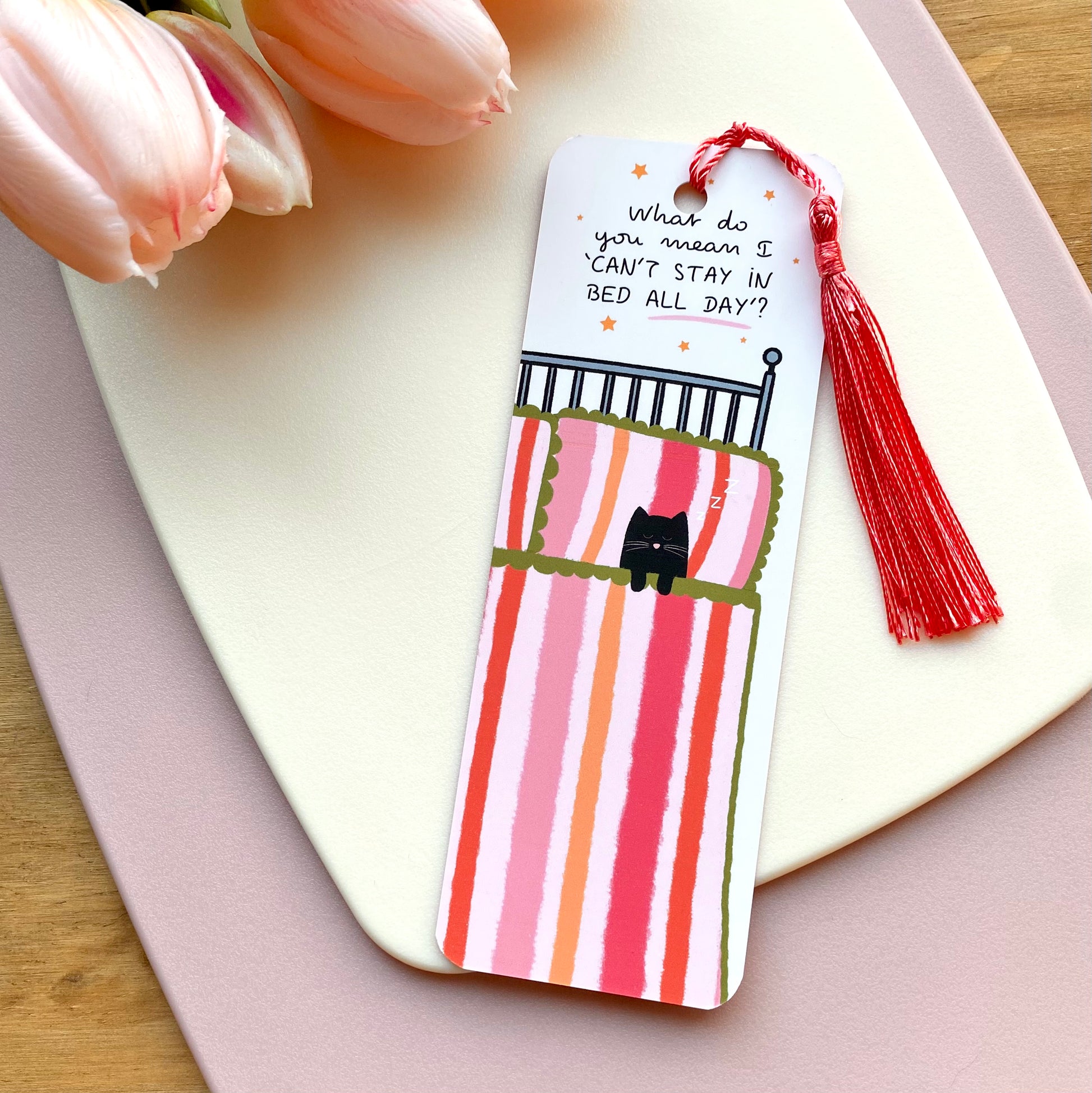 Bookmark with cute cat in bed illustration with pink tassel on a cream surface with tulips