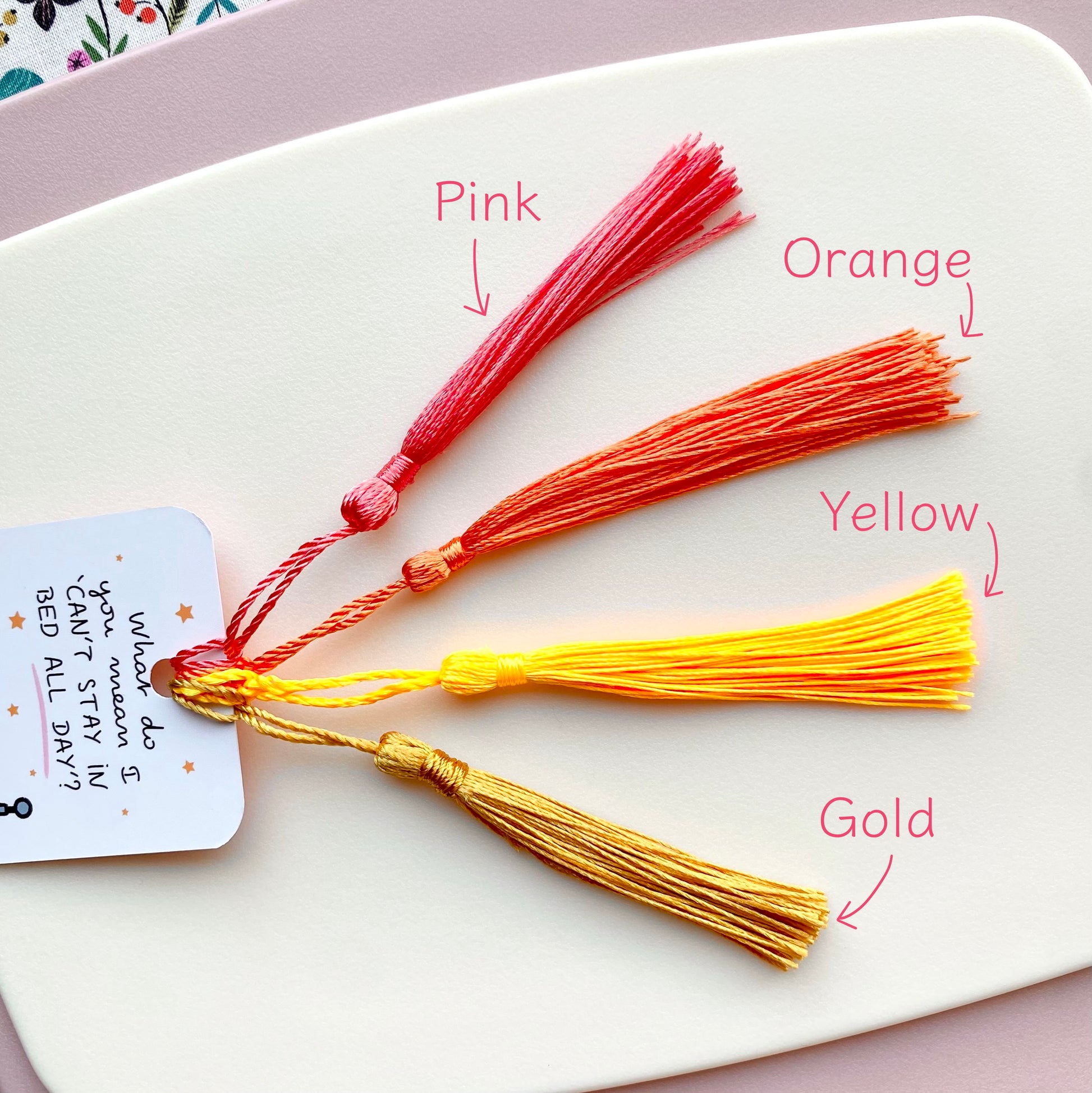 Four colourful tassels (pink, orange, yellow, gold) on a white surface with labels.