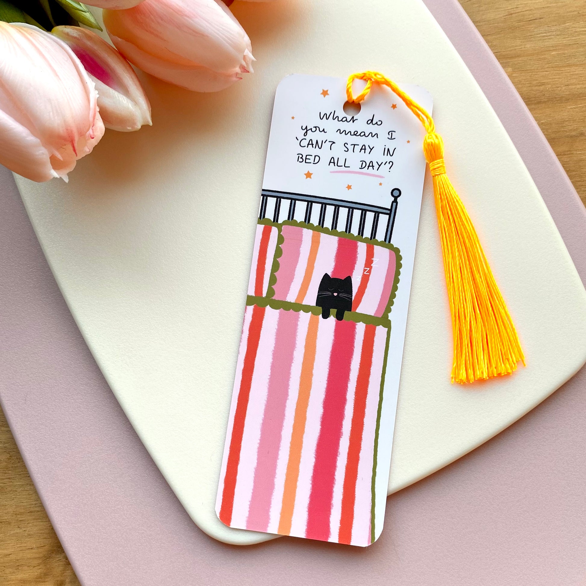 Bookmark with cute cat in bed illustration and bright yellow tassel on a pink surface with tulips