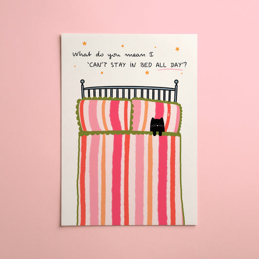 A vertical art print with a whimsical illustration of a bed with striped pink, red, and orange bedding, a black cat on the duvet, and handwritten text reading, 'What do you mean I CAN'T STAY IN BED ALL DAY?' The card is displayed against a solid light pink background.
