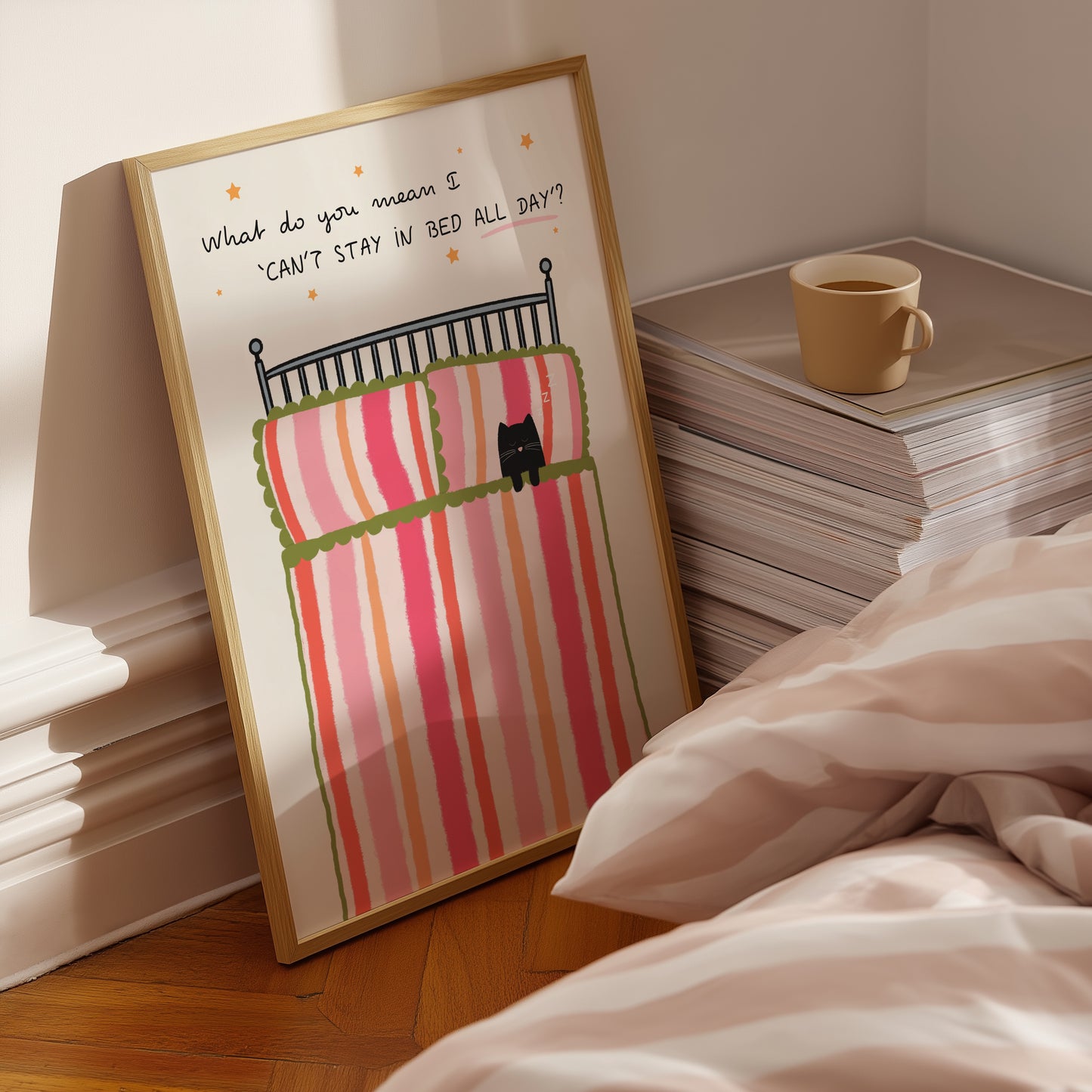 Framed girly pink striped artwork of a cat in bed with text next to a stack of books and a mug.