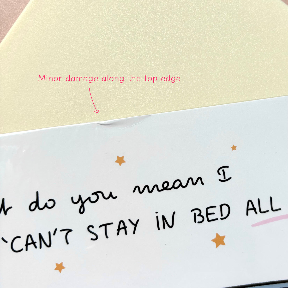 A close-up of a white illustration print with handwritten black text reading 'What do you mean I CAN'T STAY IN BED ALL DAY?' and small gold star accents. Pink annotation text says 'Minor damage along the top edge' with an arrow pointing to a crease.