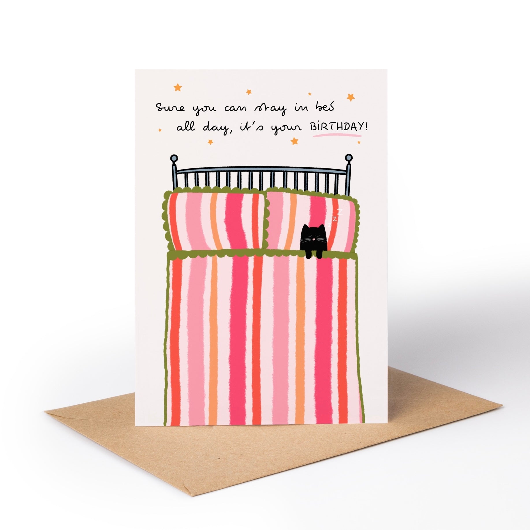 A birthday greeting card with a brown kraft envelope, featuring a whimsical illustration of a striped bed with a small black cat and the text, 'Sure you can stay in bed all day, it's your BIRTHDAY!'.