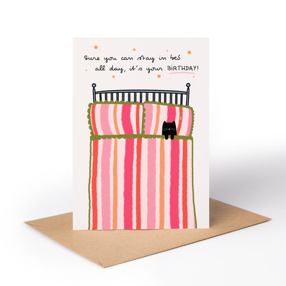A birthday greeting card with a brown kraft envelope, featuring a whimsical illustration of a striped bed with a small black cat and the text, 'Sure you can stay in bed all day, it's your BIRTHDAY!'.