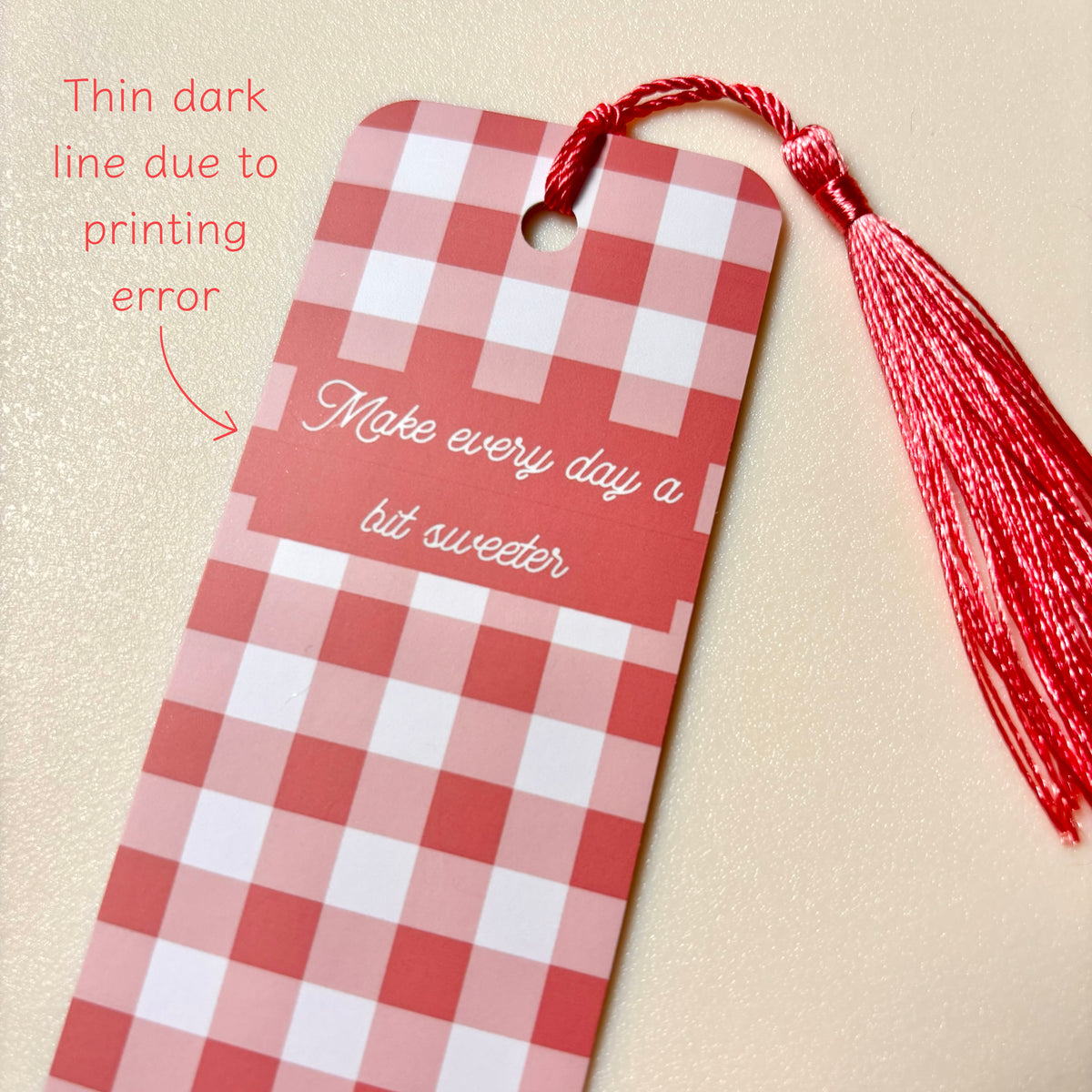 A red and white gingham bookmark with a red tassel and the phrase 'Make every day a bit sweeter' printed on it, shown on a textured beige background with a note about a thin dark line from printing.