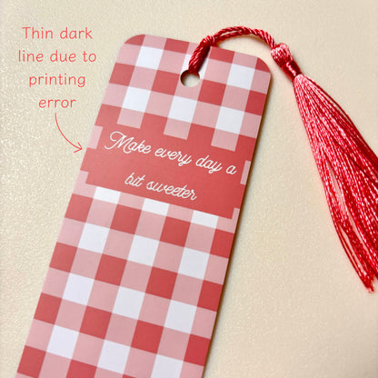 A red and white gingham bookmark with a red tassel and the phrase 'Make every day a bit sweeter' printed on it, shown on a textured beige background with a note about a thin dark line from printing.
