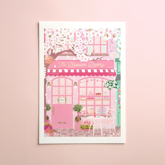 A vertical art print featuring a whimsical pink bakery storefront with cherry blossoms, displayed on a light pink wall. The print includes the text 'The Blossom Bakery' and 'The Rosem Bakery' on the facade, with pink, white, and soft green tones throughout.