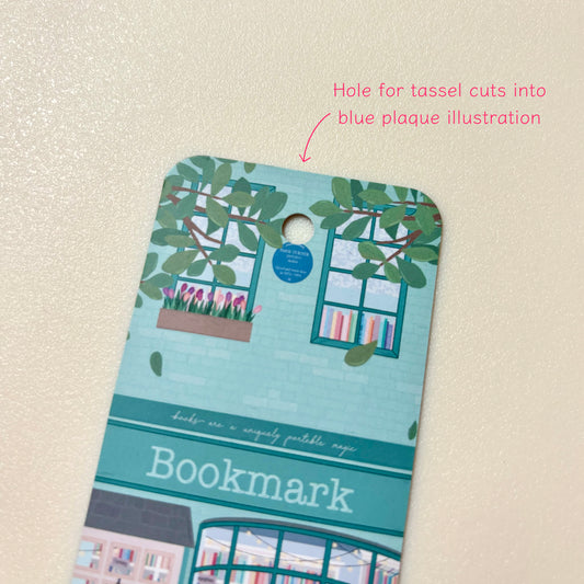 A decorative bookmark with a light blue and teal illustration of a bookstore facade, featuring windows, green leaves, pink tulips, and the word 'Bookmark' on a textured cream background. Pink text at the top right notes the tassel hole cuts into the illustration slightly.