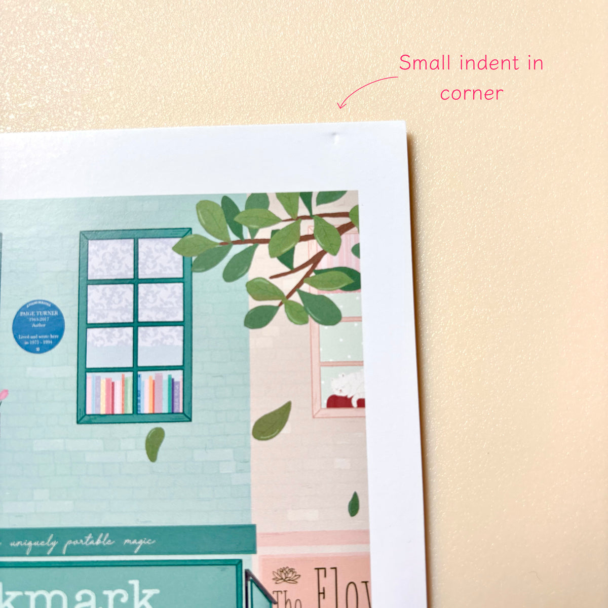 A close-up of a print showing part of an illustrated bookshop scene with green leaves, a window, and pastel colours, placed on a beige textured background. Pink text at the top reads 'Small indent in corner'.