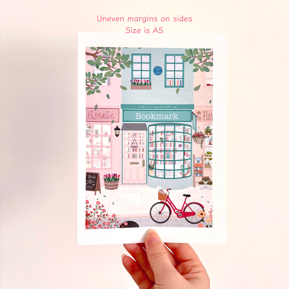 A hand holds an A5 size art print featuring a whimsical illustration of a bookshop storefront with pink and teal details, a pink bicycle, and floral accents. Pink text at the top reads 'Uneven margins on sides' and 'Size is A5'.