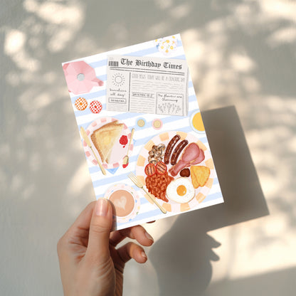 Hand holding an A6 card with breakfast food illustrations and 'The Birthday Times' text on a newspaper.