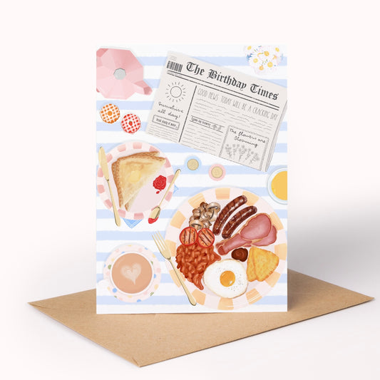 A birthday greeting card featuring a breakfast illustration with a newspaper headline, toast, beans, sausage, egg, and coffee, displayed upright on a brown envelope against a white background.